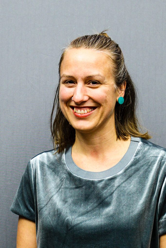 Leende kvinna med rakt brunt hår, blågröna örhängen och en grå topp, mot en grå bakgrund.