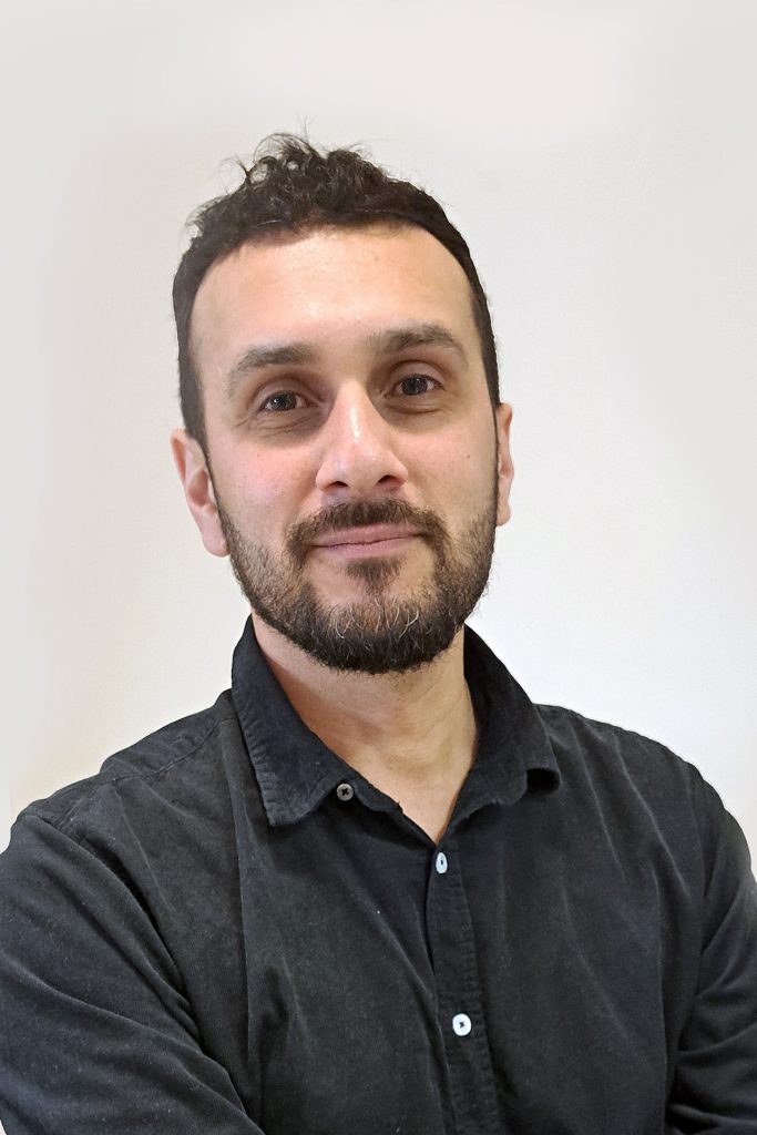 Man med kort mörkt hår och skägg, klädd i svart skjorta med knappar, ler lätt mot en enfärgad bakgrund.