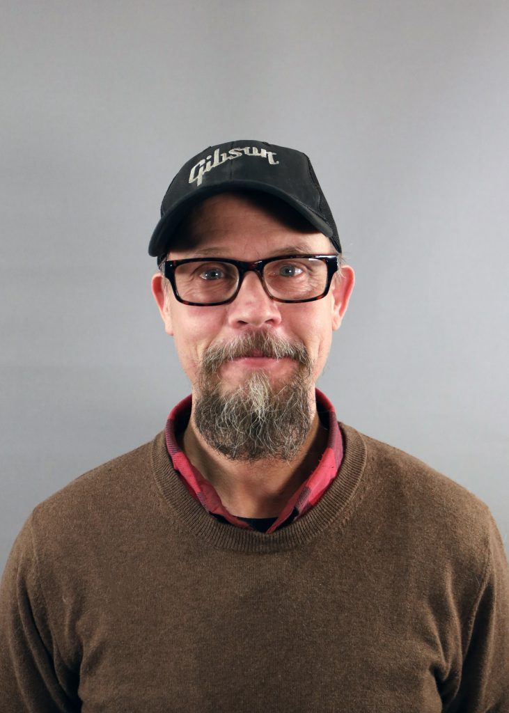 Man med glasögon, skägg och svart Gibsonkeps, klädd i brun tröja över röd skjorta med krage.