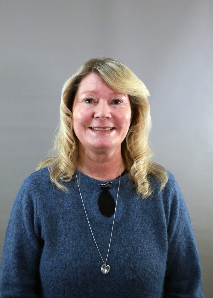 Leende kvinna med blont hår, iförd blå tröja och hängande halsband, mot grå bakgrund.