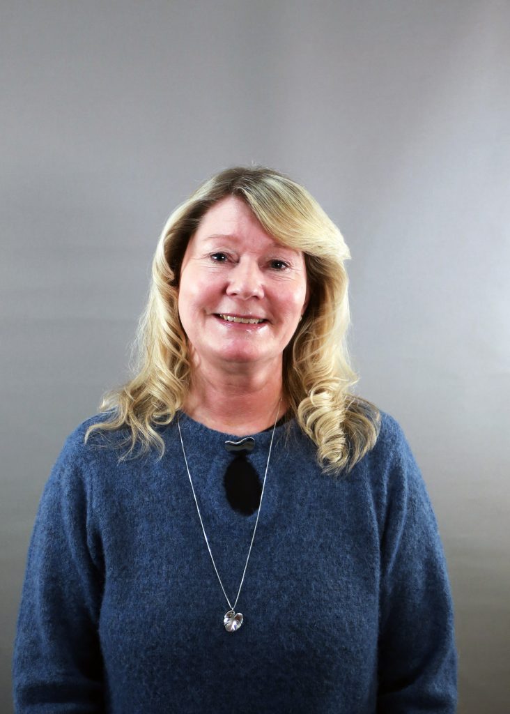 En kvinna med blont hår, blå tröja och halsband ler mot en grå bakgrund.