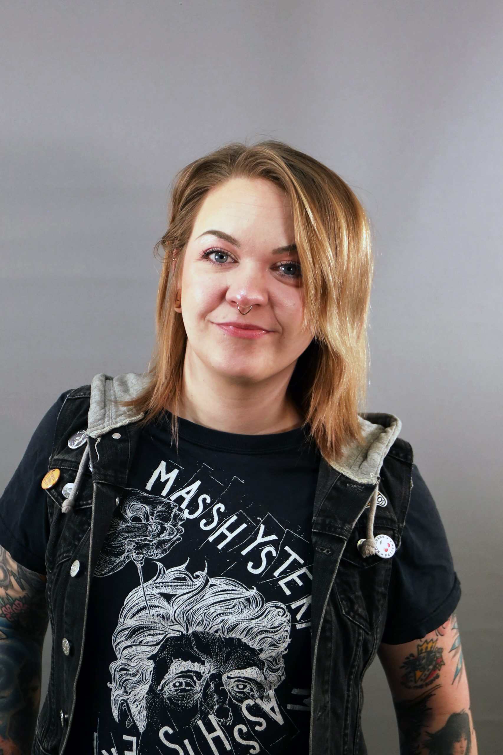 Leende person med tatueringar iförd en grafisk t-shirt och en svart väst, stående mot en grå bakgrund.