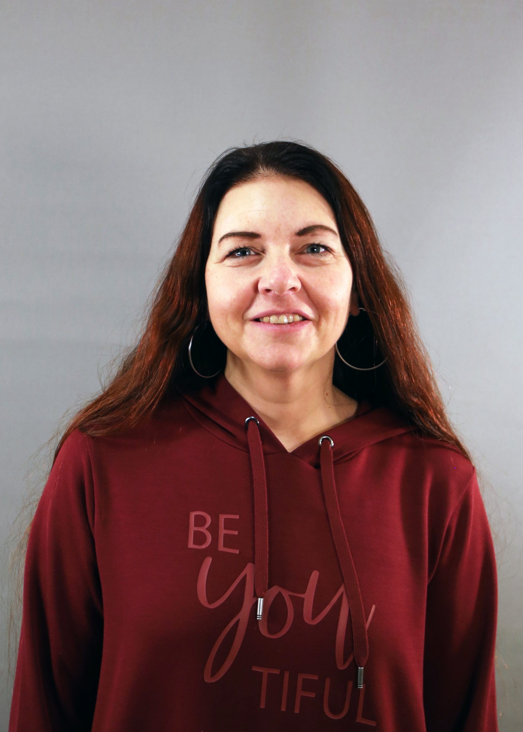 Kvinna med långt brunt hår i rödbrun huvtröja med texten "Be You Tiful" ler mot en grå bakgrund.