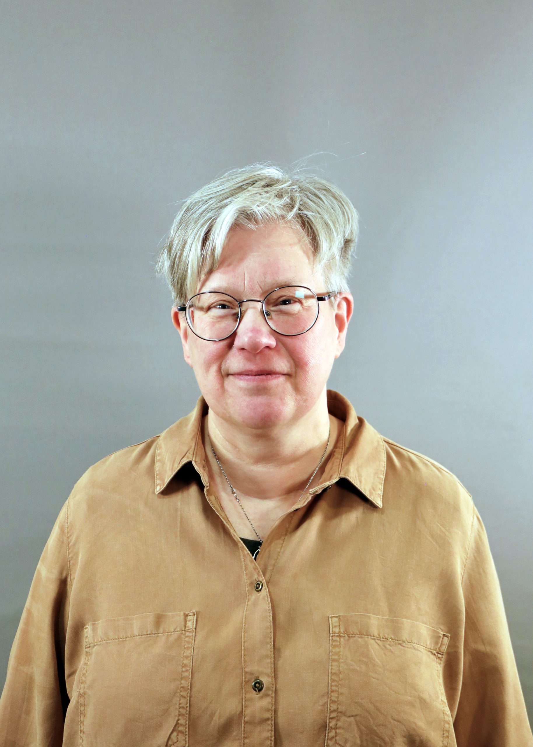 Person med kort grått hår och glasögon, klädd i brun skjorta med knappar, står mot en enfärgad bakgrund.