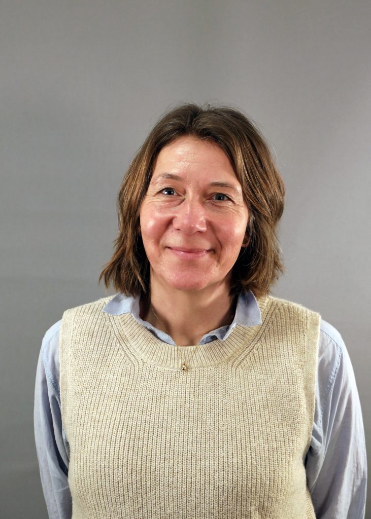 En kvinna med brunt hår, klädd i en beige tröja över en ljusblå skjorta, ler mot kameran.