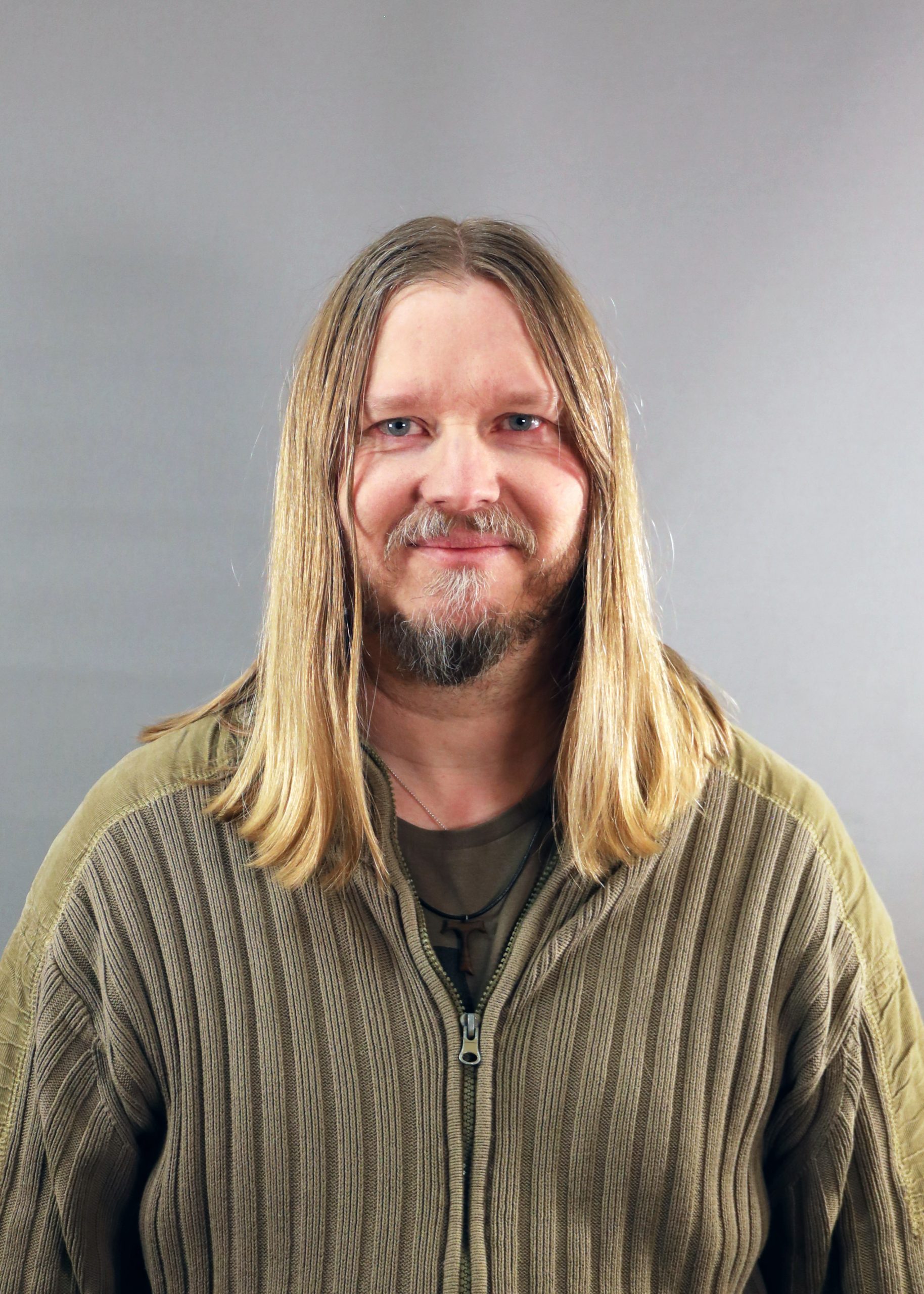 En man med långt blont hår och skägg ler, iklädd en brun tröja med dragkedja, mot en grå bakgrund.