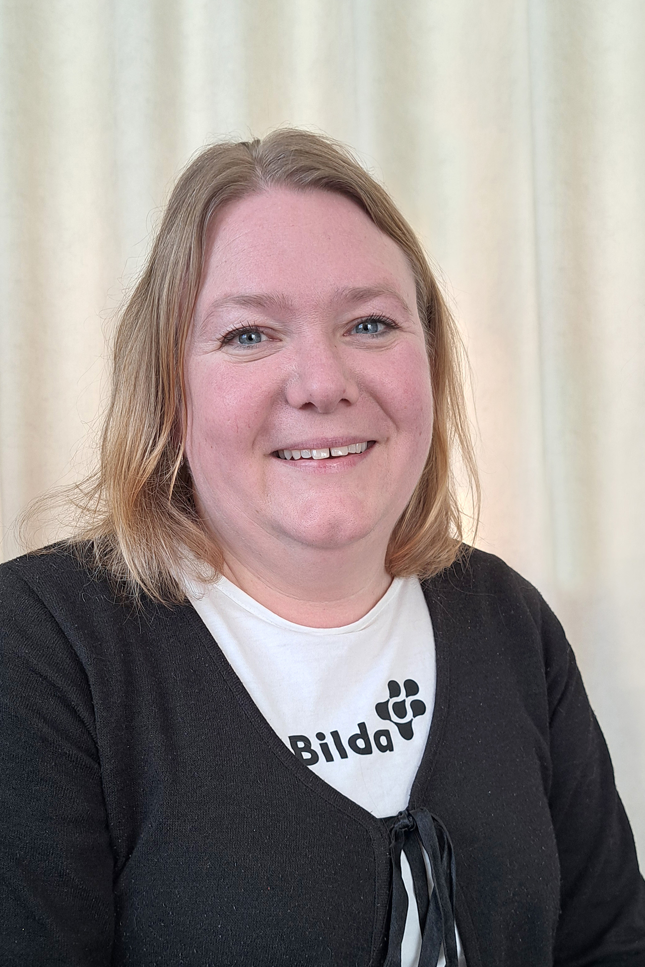En leende kvinna med axellångt blont hår, klädd i vit skjorta och svart kofta, framför en ljusridå.