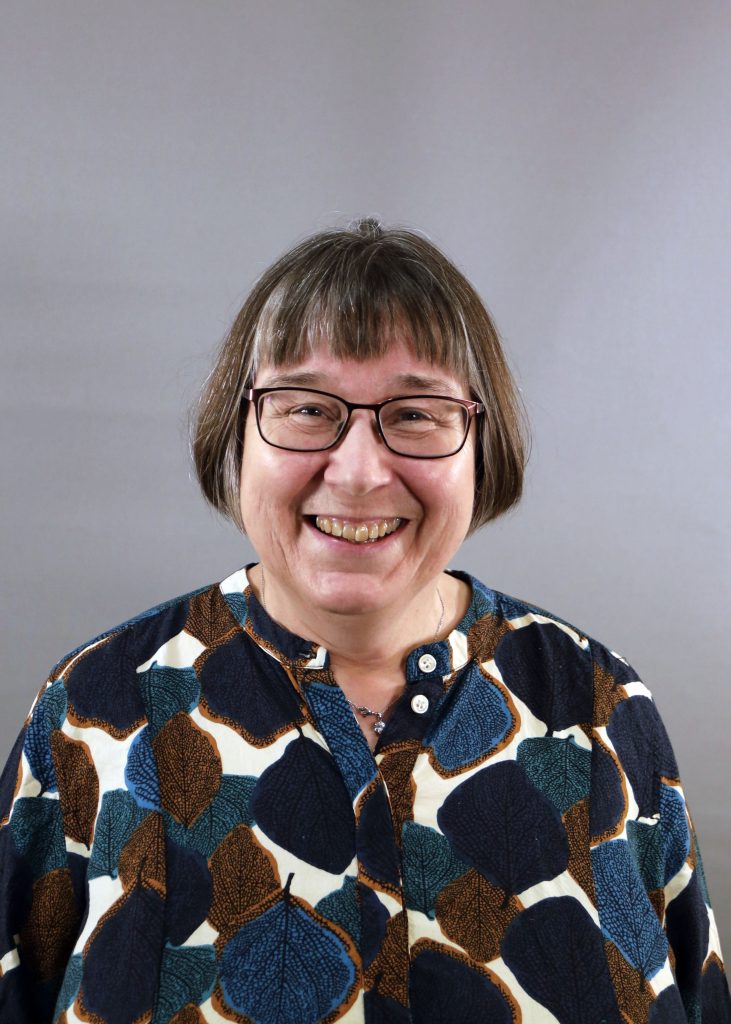 En leende kvinna med glasögon och kort hår, klädd i mönstrad skjorta, mot en enfärgad grå bakgrund.