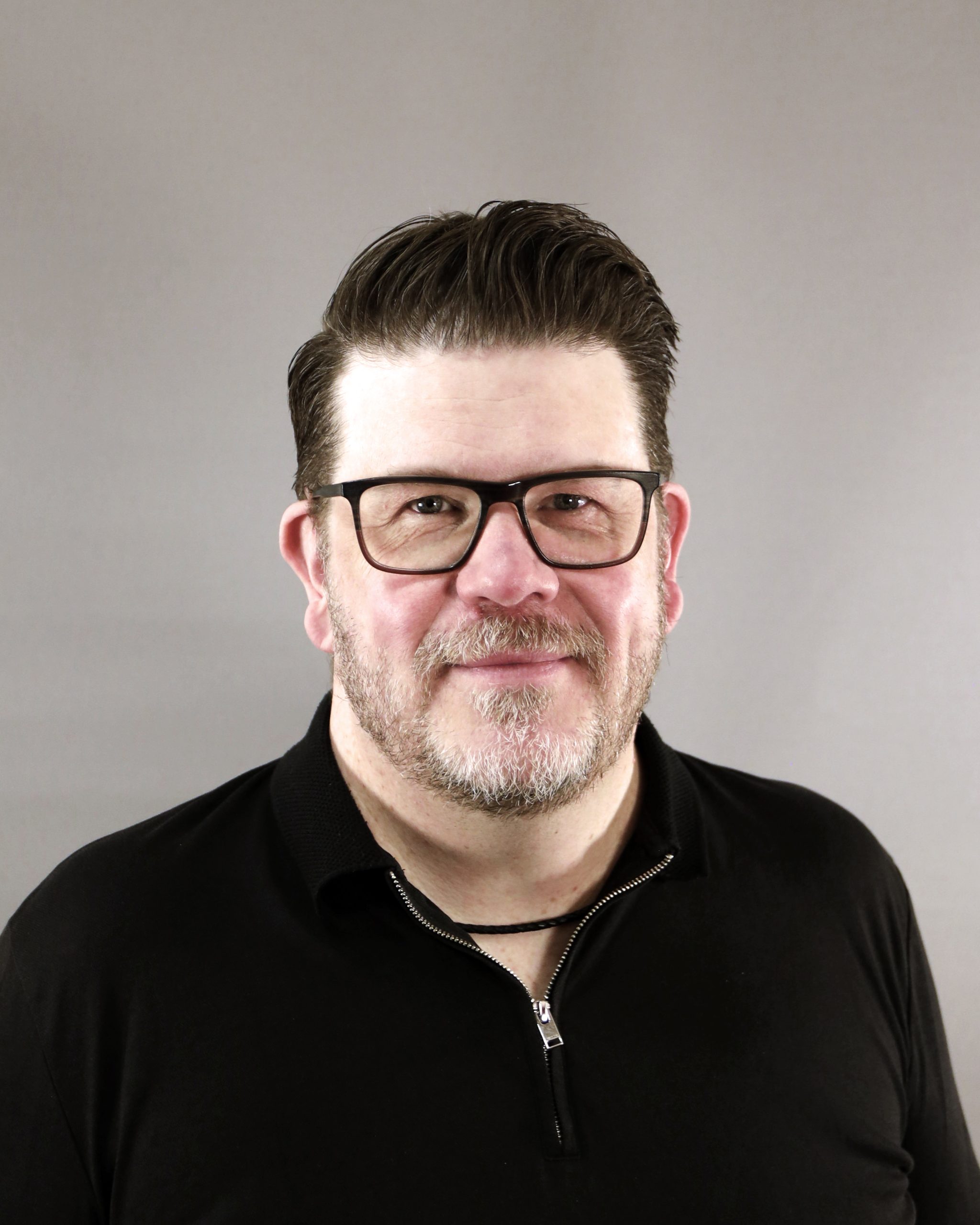 Leende man med glasögon och skägg iklädd svart skjorta mot en enfärgad grå bakgrund.