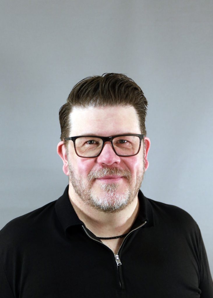 En man med glasögon och skägg ler lätt, klädd i svart skjorta mot en grå bakgrund.