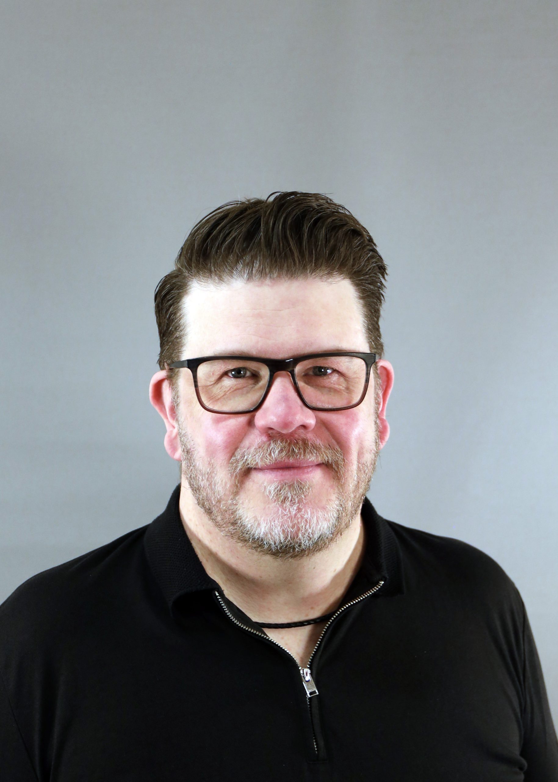 En man med glasögon och skägg ler lätt, klädd i svart skjorta mot en grå bakgrund.