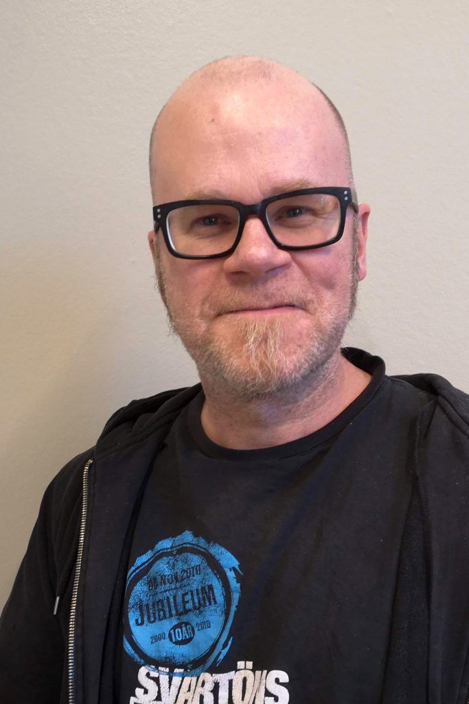 En man med glasögon och kort skägg, klädd i svart T-shirt och huvtröja, ler mot kameran.