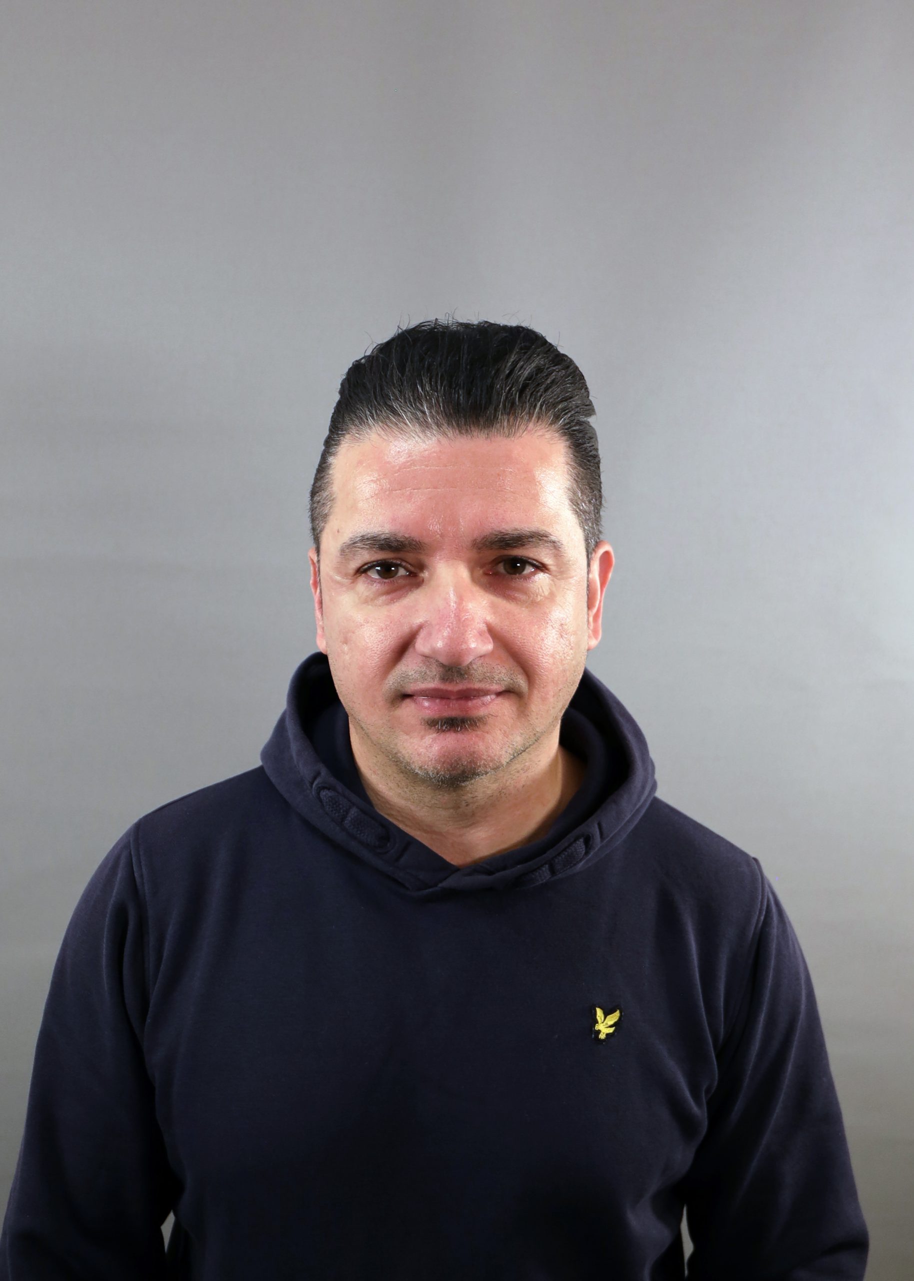 Man med kort mörkt hår i mörk huvtröja står mot en grå bakgrund och vänder sig mot kameran.