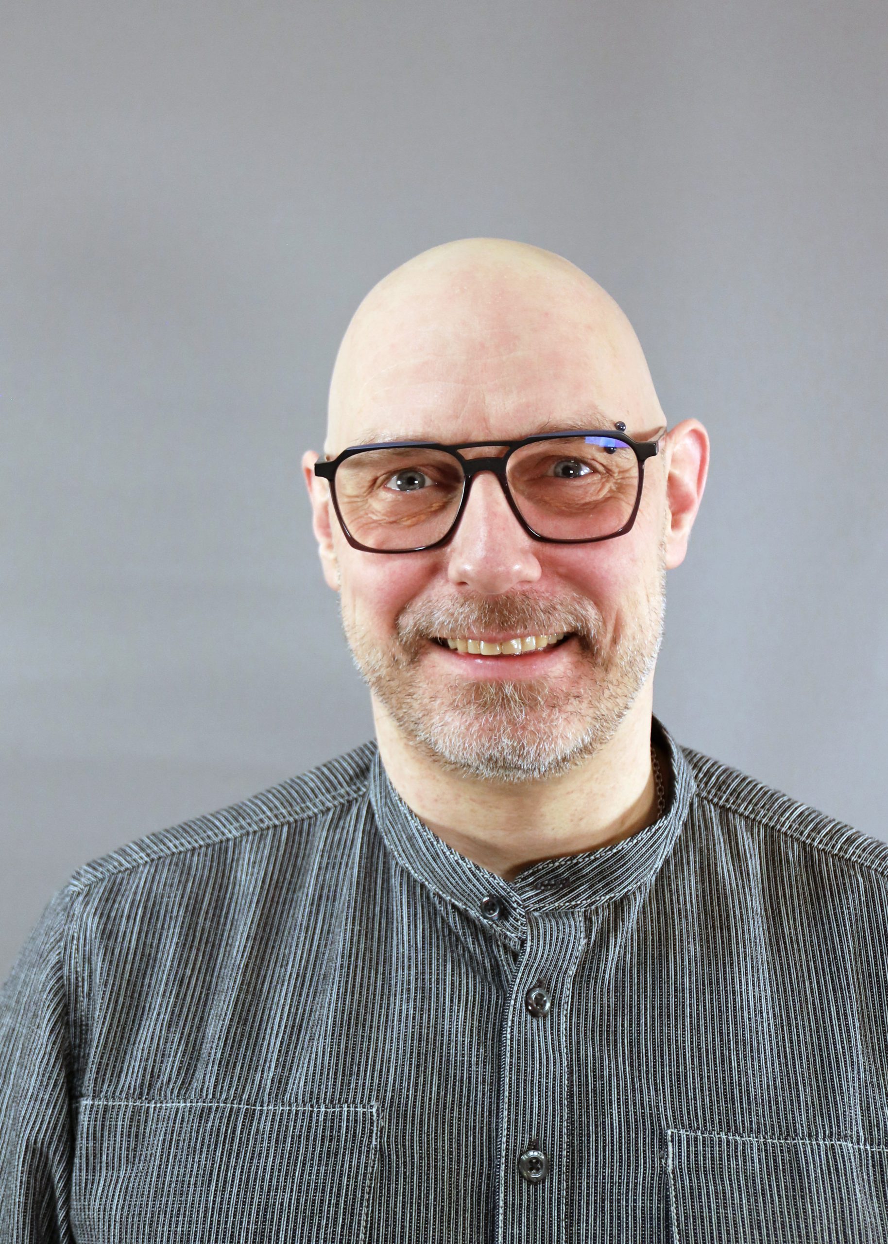 En leende skallig man med glasögon och randig skjorta står framför en grå bakgrund.