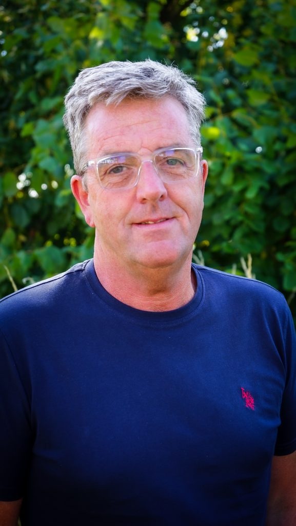 En man med grått hår och glasögon, klädd i en marinblå t-shirt, står utomhus framför gröna bladverk.