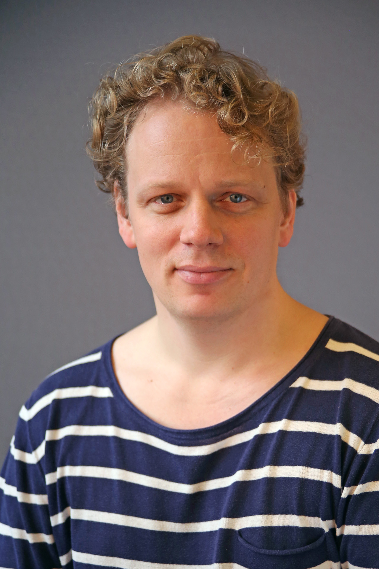 En person med lockigt blont hår, klädd i en marinblå och vit randig skjorta, ler mjukt mot kameran.