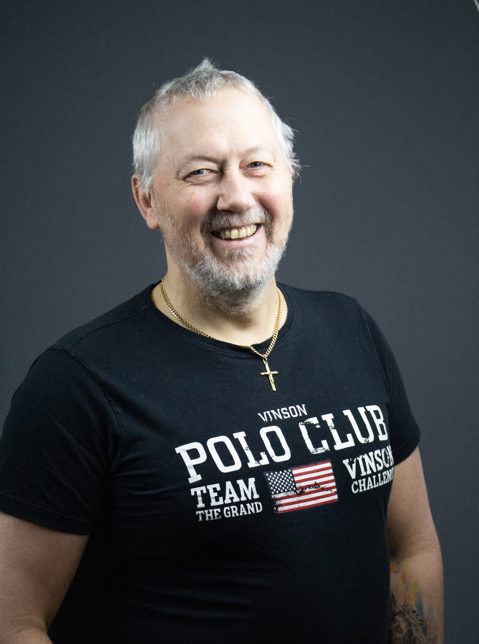 Leende man med kort grått hår och skägg, klädd i svart "Polo Club"-t-shirt och korshalsband i guld.