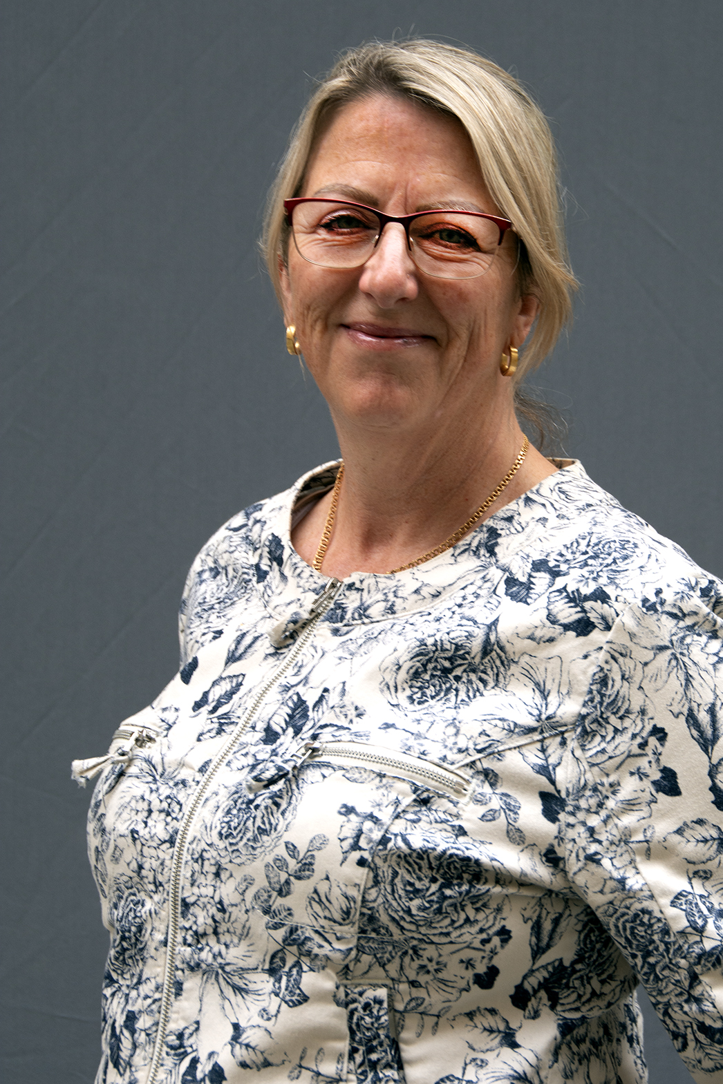 En leende kvinna med glasögon, klädd i en vit och blå jacka med blommönster, står framför en enfärgad grå bakgrund.