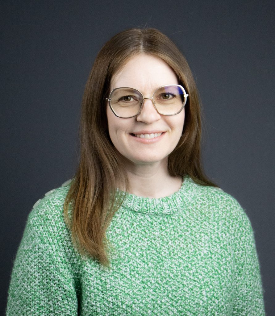 Kvinna med långt brunt hår, glasögon och grön tröja ler mot en mörkgrå bakgrund.