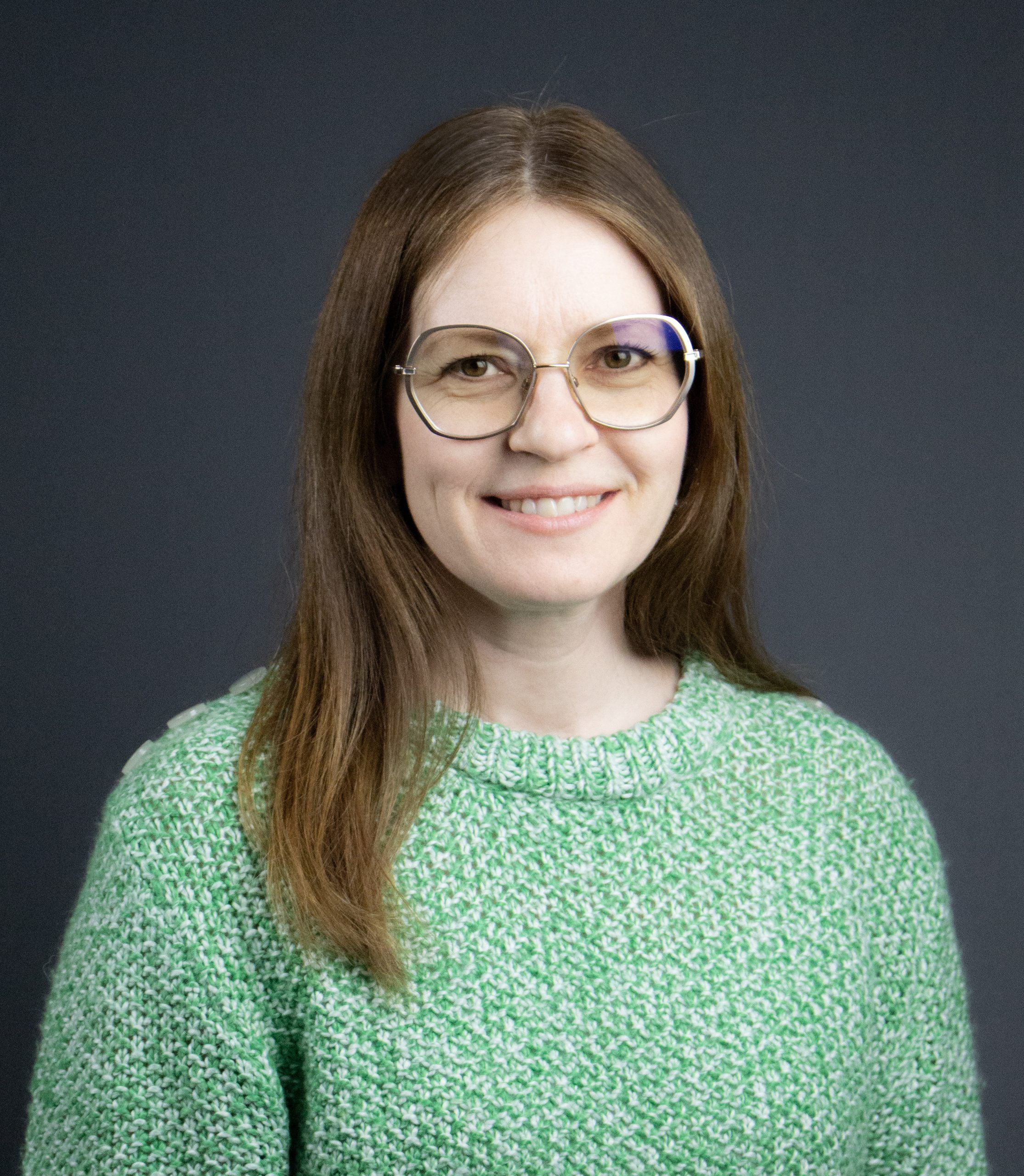 Kvinna med långt brunt hår, glasögon och grön tröja ler mot en mörkgrå bakgrund.