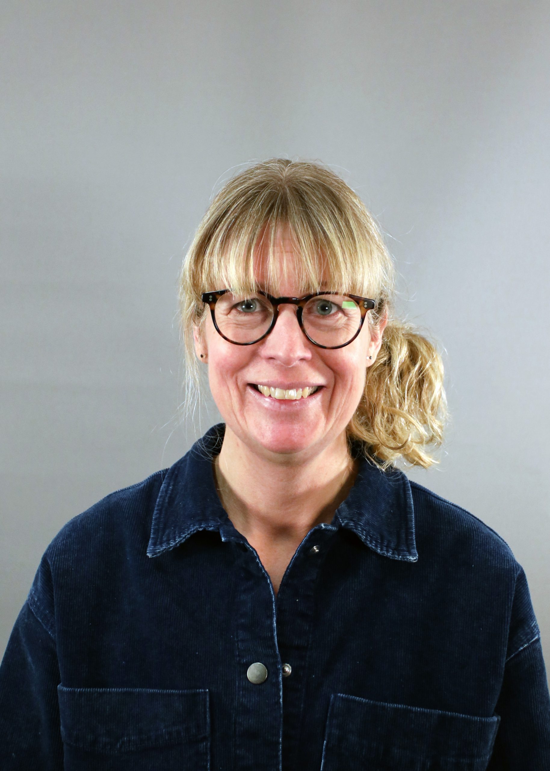 En kvinna med blont hår i hästsvans, glasögon och mörk jacka ler mot en grå bakgrund.