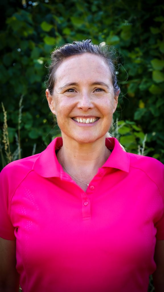 Kvinna i ljusrosa polotröja ler utomhus med gröna bladverk i bakgrunden.