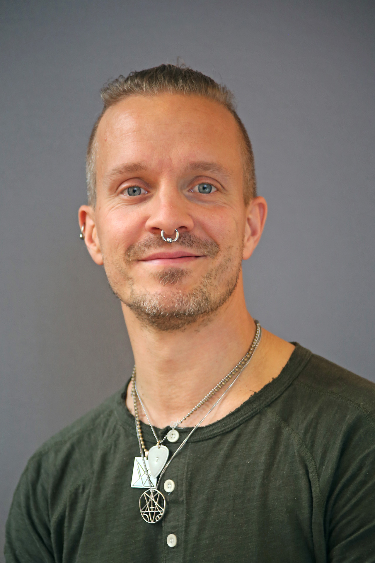 En person med kort hår, piercingar i ansiktet och halsband i flera lager ler mot en grå bakgrund.