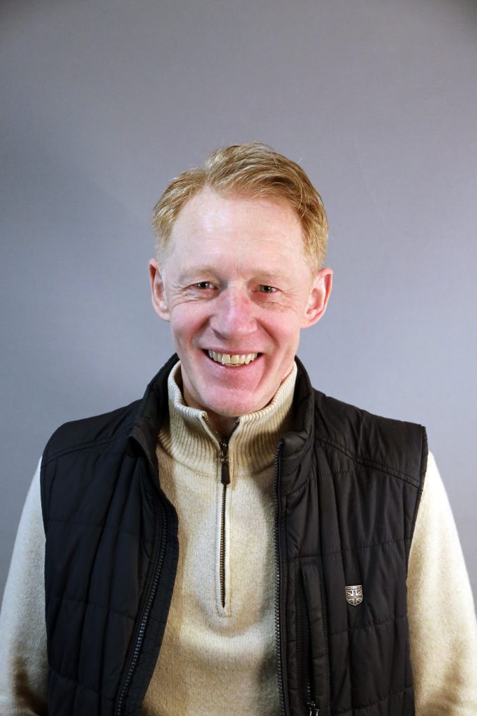 Leende man med kort ljust hår, klädd i ljus tröja och svart väst, stående mot grå bakgrund.