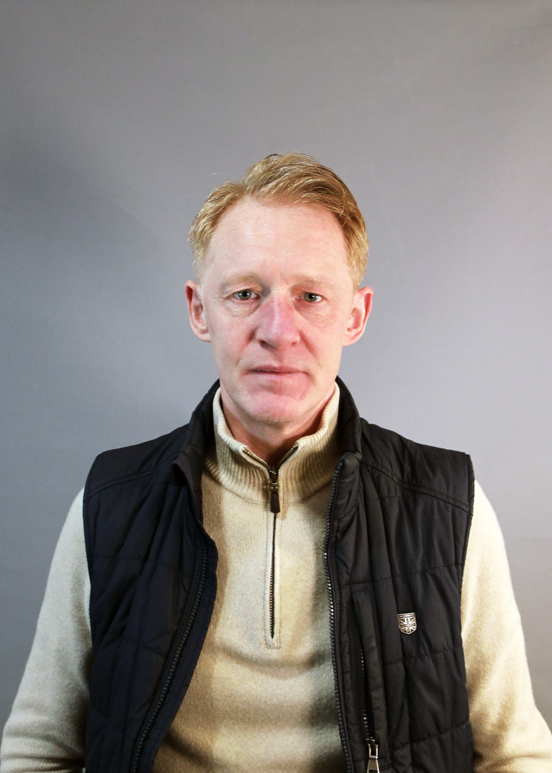 En man med ljust hår i beige tröja och svart väst står mot en grå bakgrund.