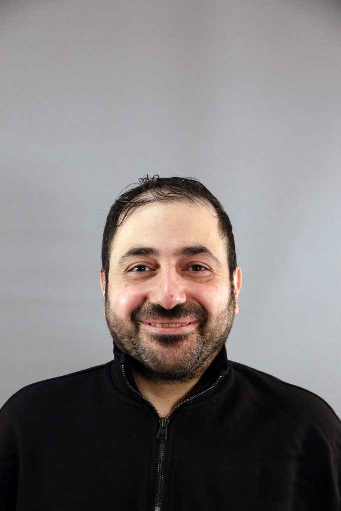 En man med kort mörkt hår och skägg ler mot kameran mot en grå bakgrund.