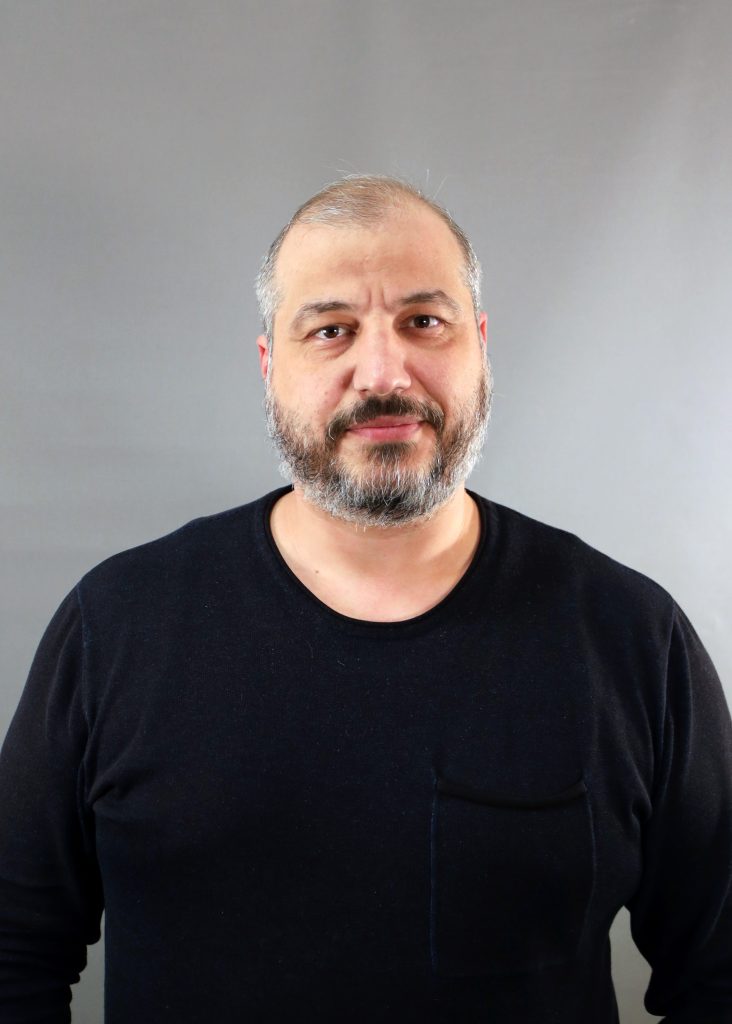 En man med skägg och kort hår, klädd i svart skjorta, står mot en grå bakgrund.