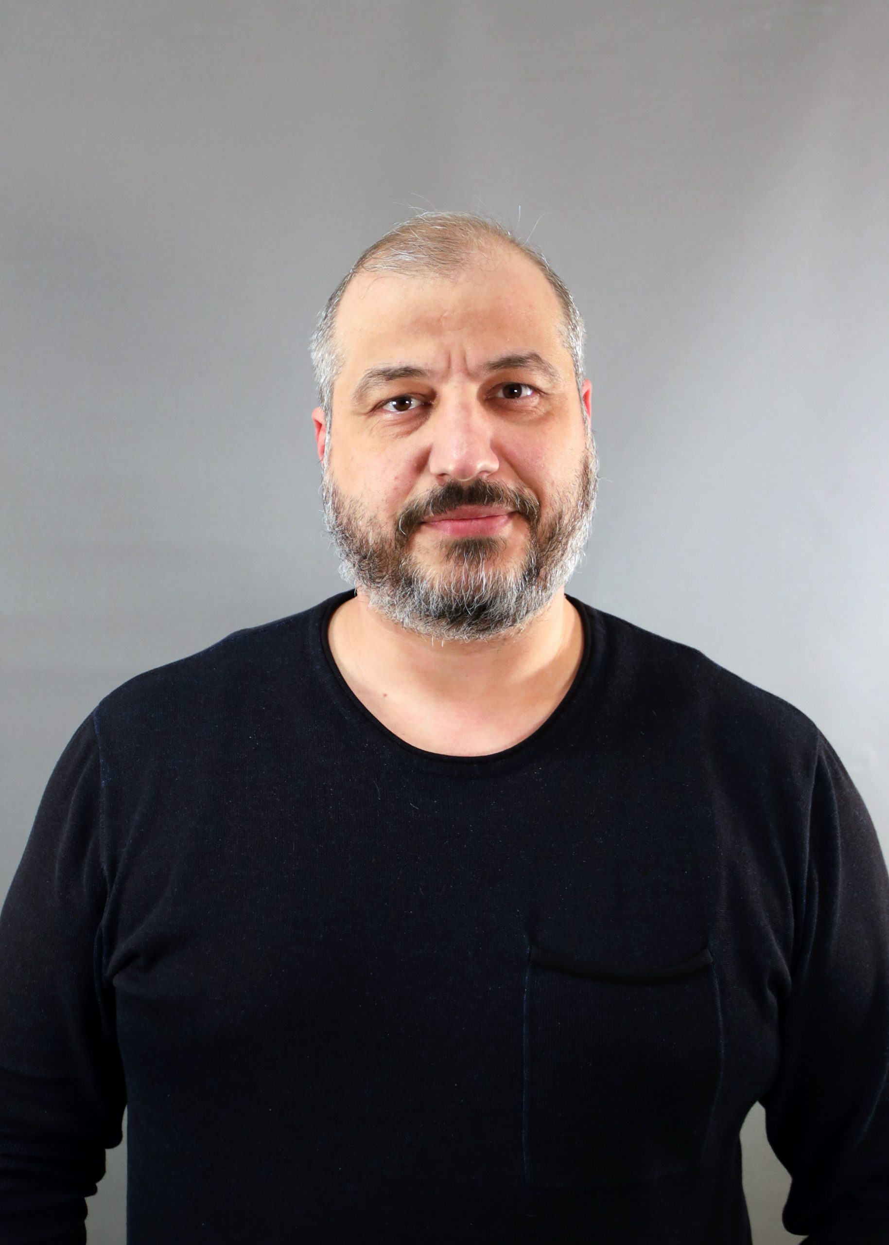 En man med skägg och kort hår, klädd i svart skjorta, står mot en grå bakgrund.