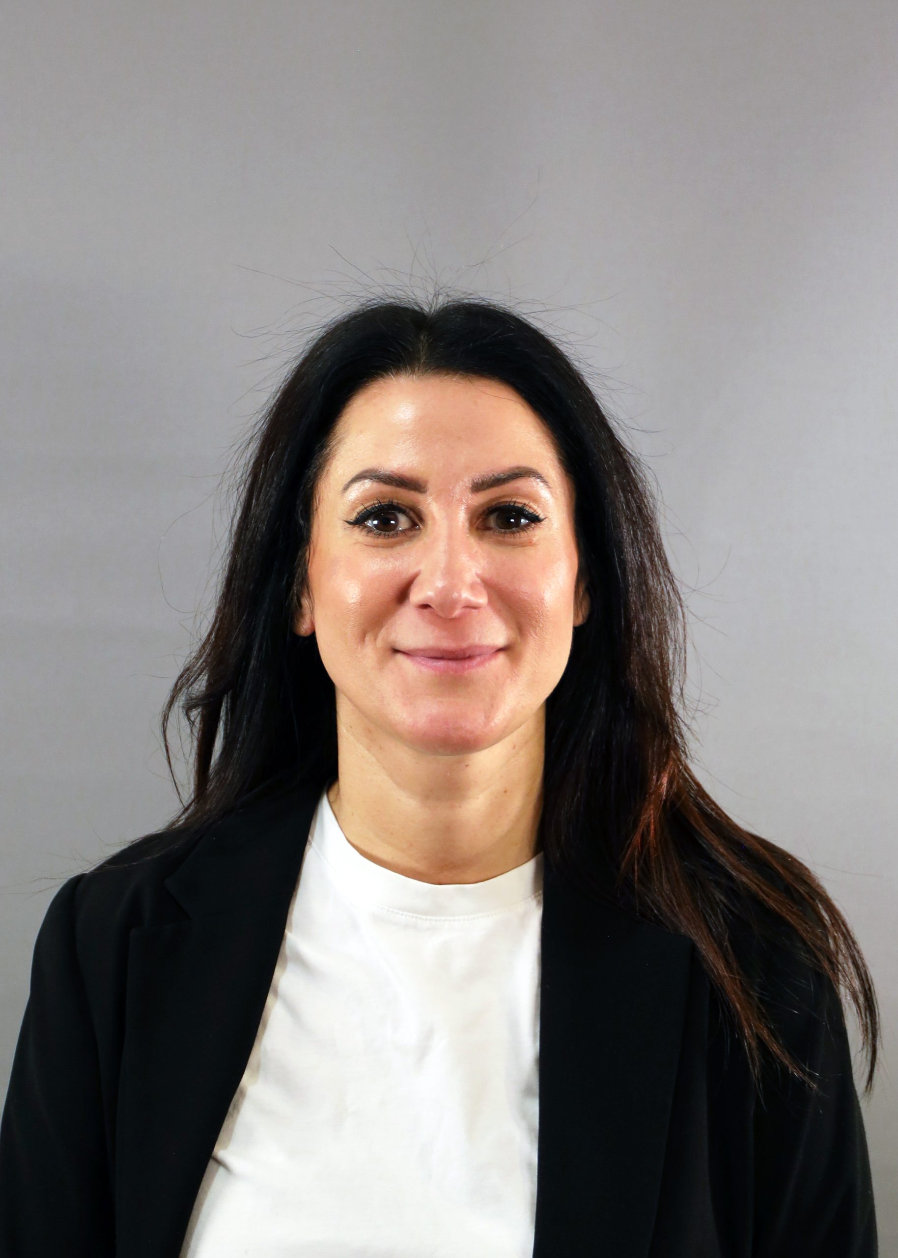 Kvinna med långt mörkt hår i svart kavaj och vit skjorta, ler mot en grå bakgrund.