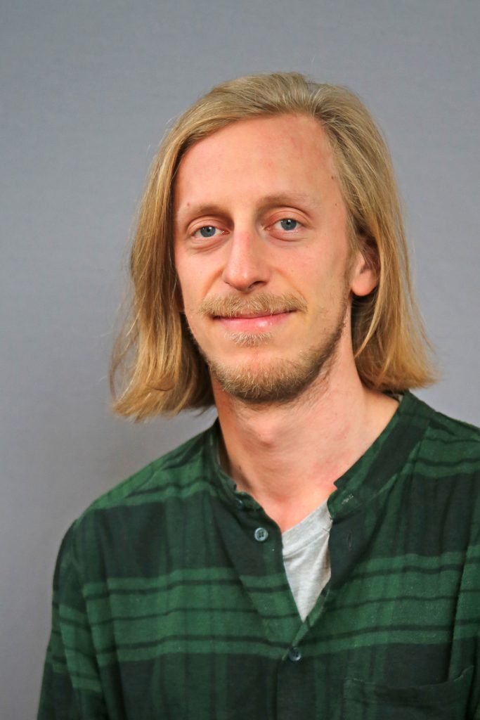 En man med axellångt blont hår och en grön rutig skjorta ler mot kameran mot en grå bakgrund.