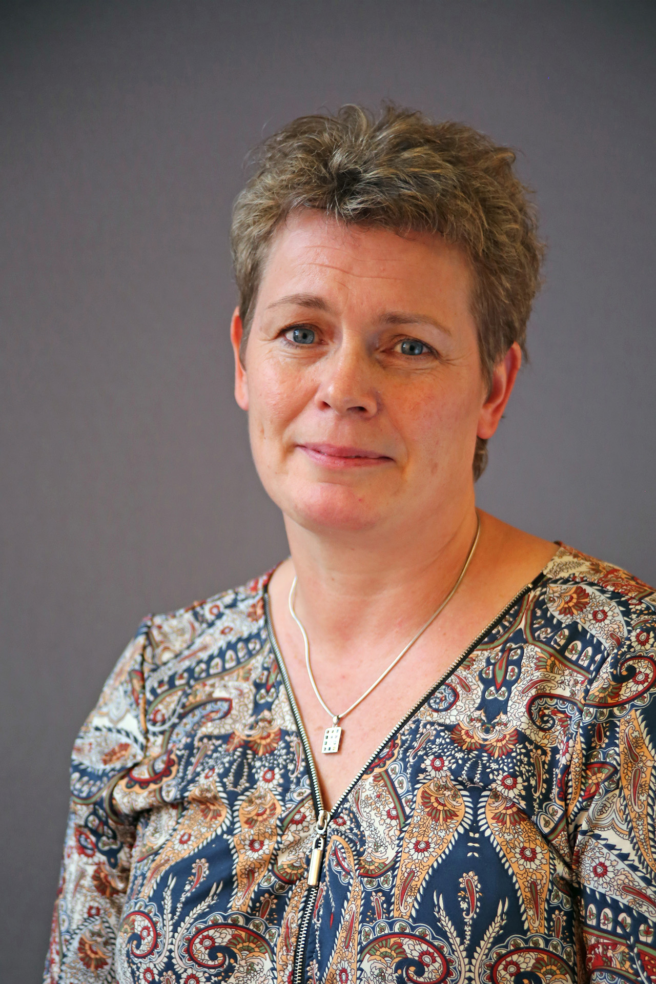 En kvinna med kort hår i mönstrad blus och halsband står mot en enfärgad bakgrund.