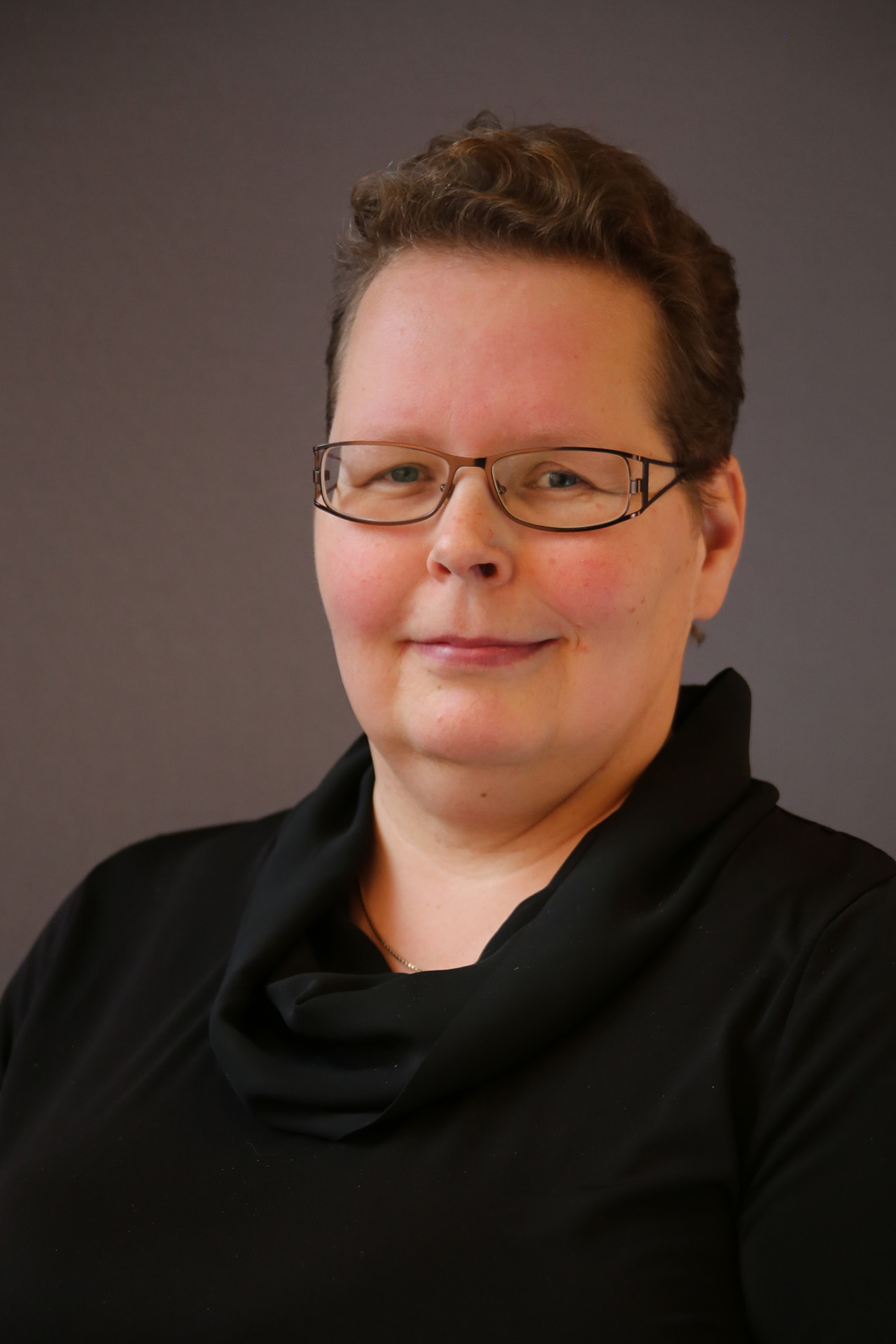 En person med kort hår och glasögon, klädd i svart topp, poserar mot en vanlig, mörk bakgrund.