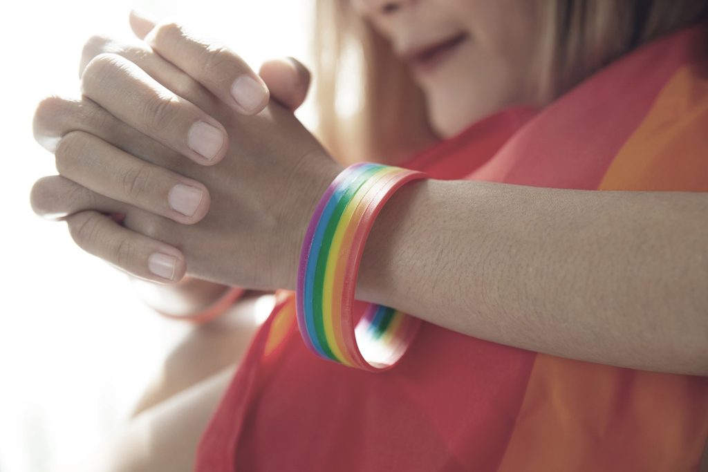 Person med knäppta händer, bär ett regnbågspride-armband och är draperad i en regnbågsflagga.