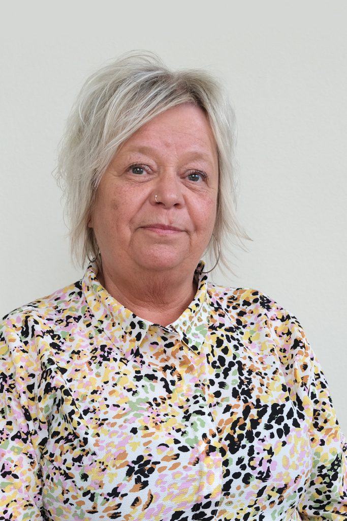 En kvinna med kort, ljust hår bär en färgstark mönstrad skjorta mot en enfärgad bakgrund.