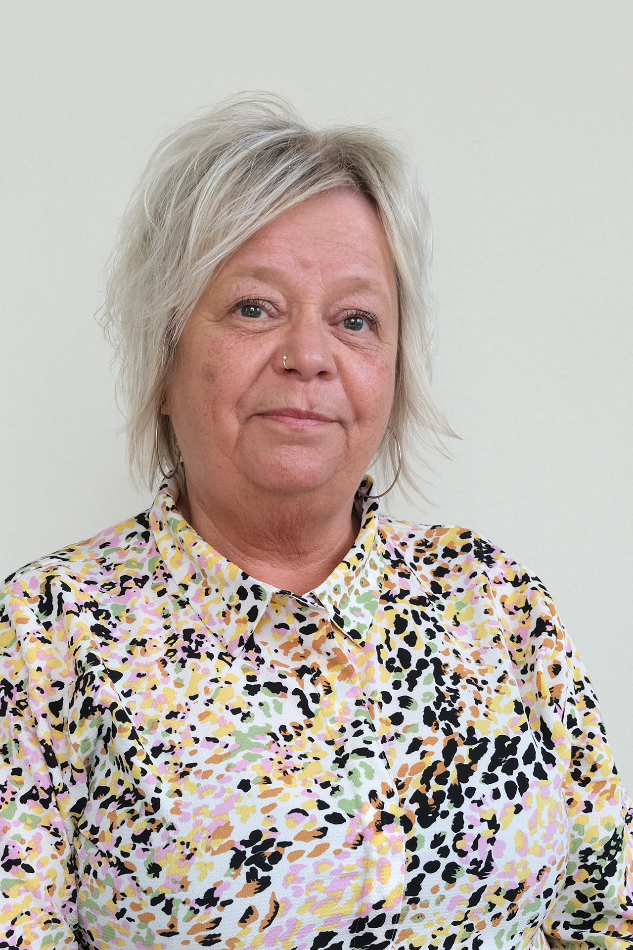 En kvinna med kort, ljust hår bär en färgstark mönstrad skjorta mot en enfärgad bakgrund.