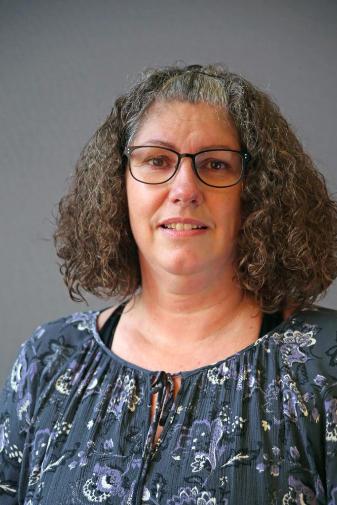 En kvinna med lockigt hår och glasögon, klädd i mönstrad blus, står framför en enfärgad bakgrund.