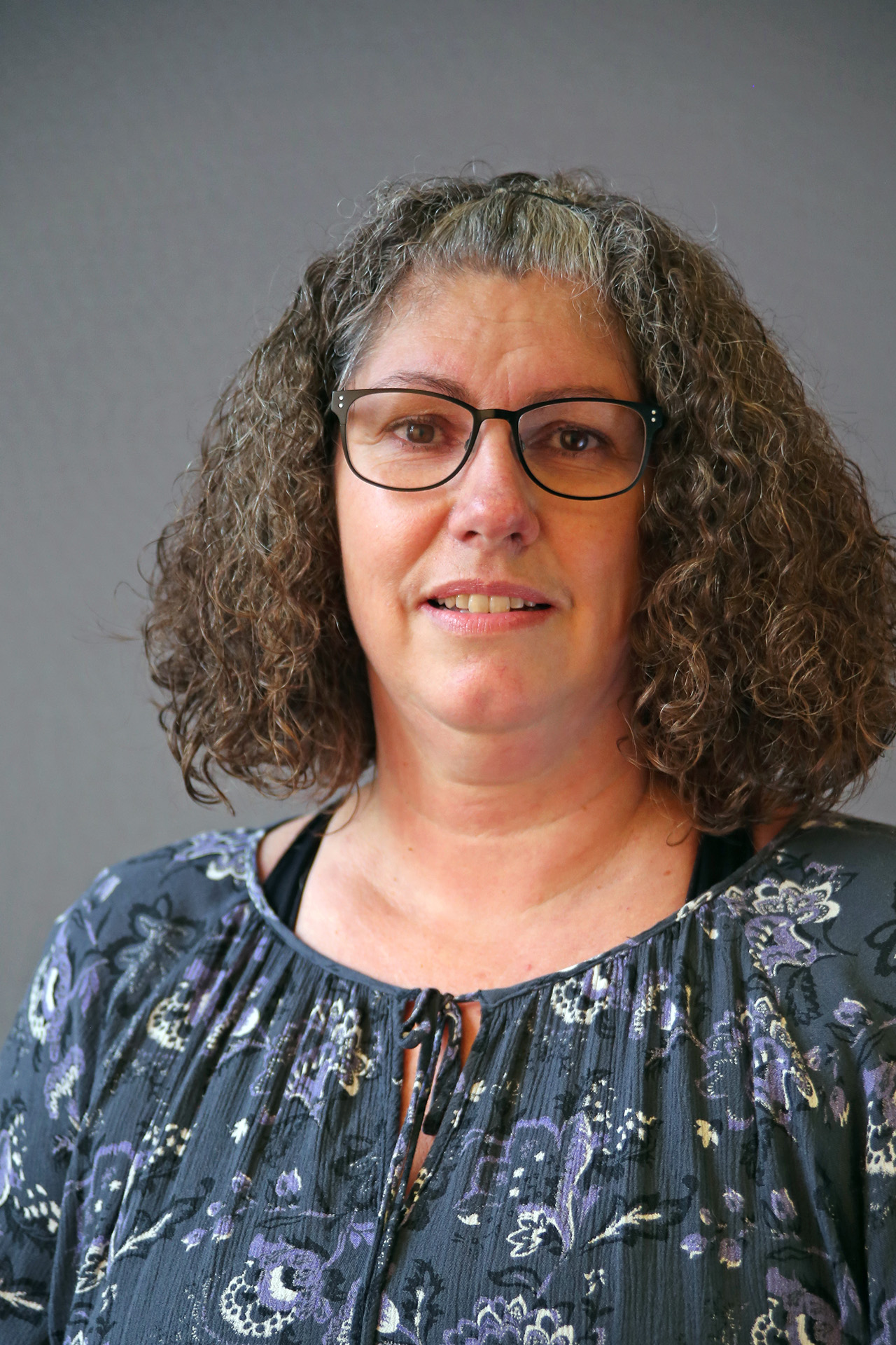 En kvinna med lockigt hår och glasögon, klädd i mönstrad blus, står framför en enfärgad bakgrund.