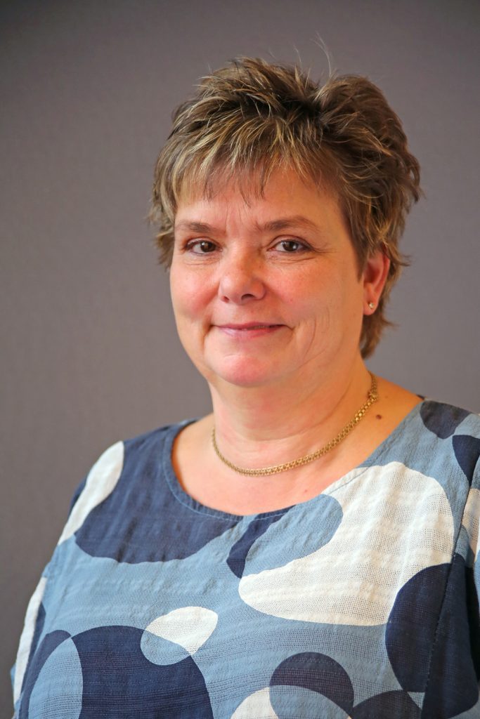 En kvinna med kort hår ler lätt, klädd i en blåmönstrad topp och ett guldhalsband mot en grå bakgrund.