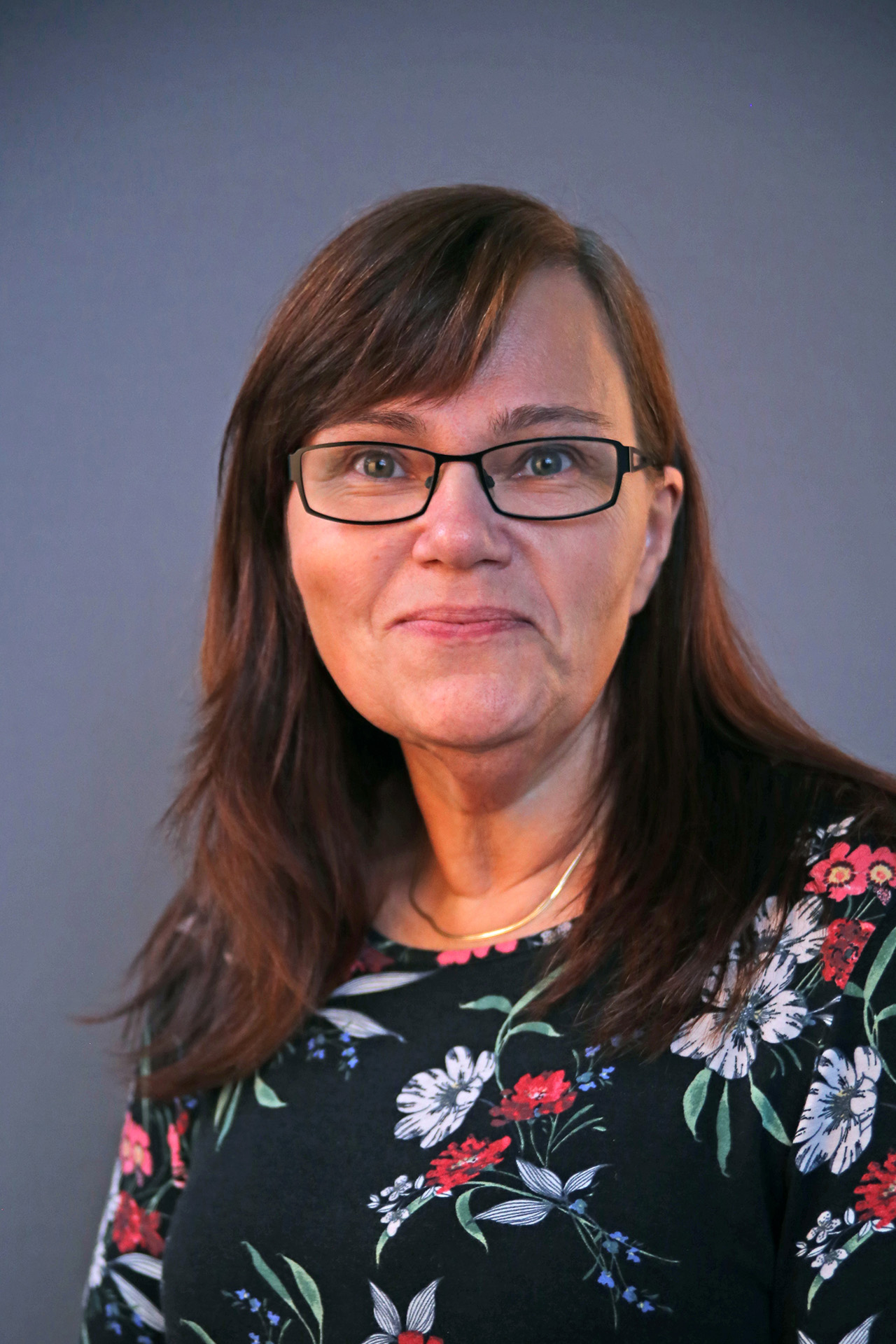 En kvinna med glasögon och långt brunt hår ler, klädd i en blommig topp, mot en enfärgad bakgrund.