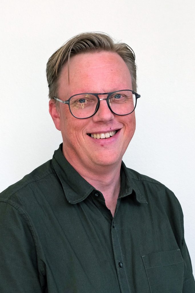 Man med glasögon och mörkgrön skjorta ler mot en vit bakgrund.