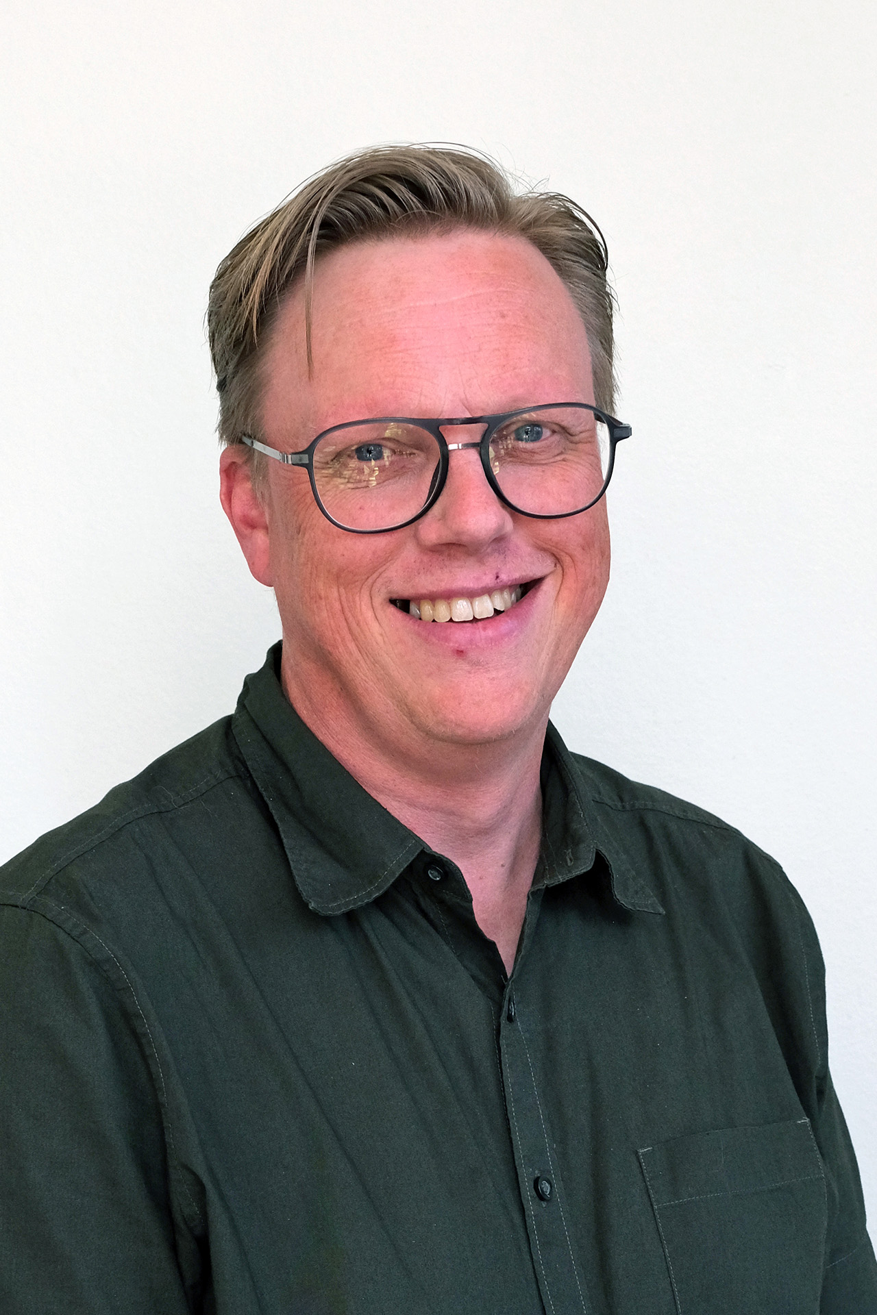 Man med glasögon och mörkgrön skjorta ler mot en vit bakgrund.