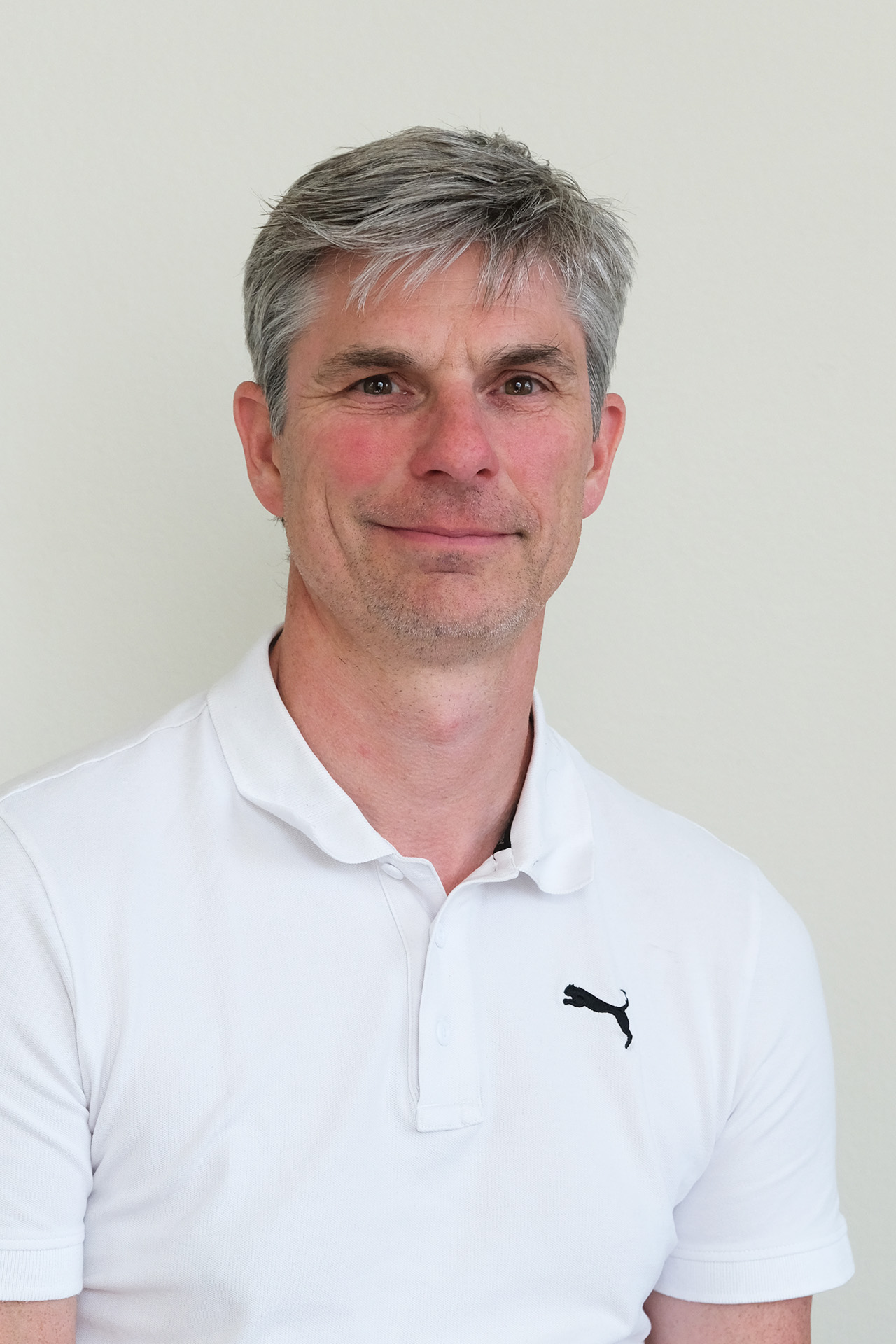 Leende man med kort grått hår i vit polotröja med svart Puma-logotyp, mot en enfärgad bakgrund.