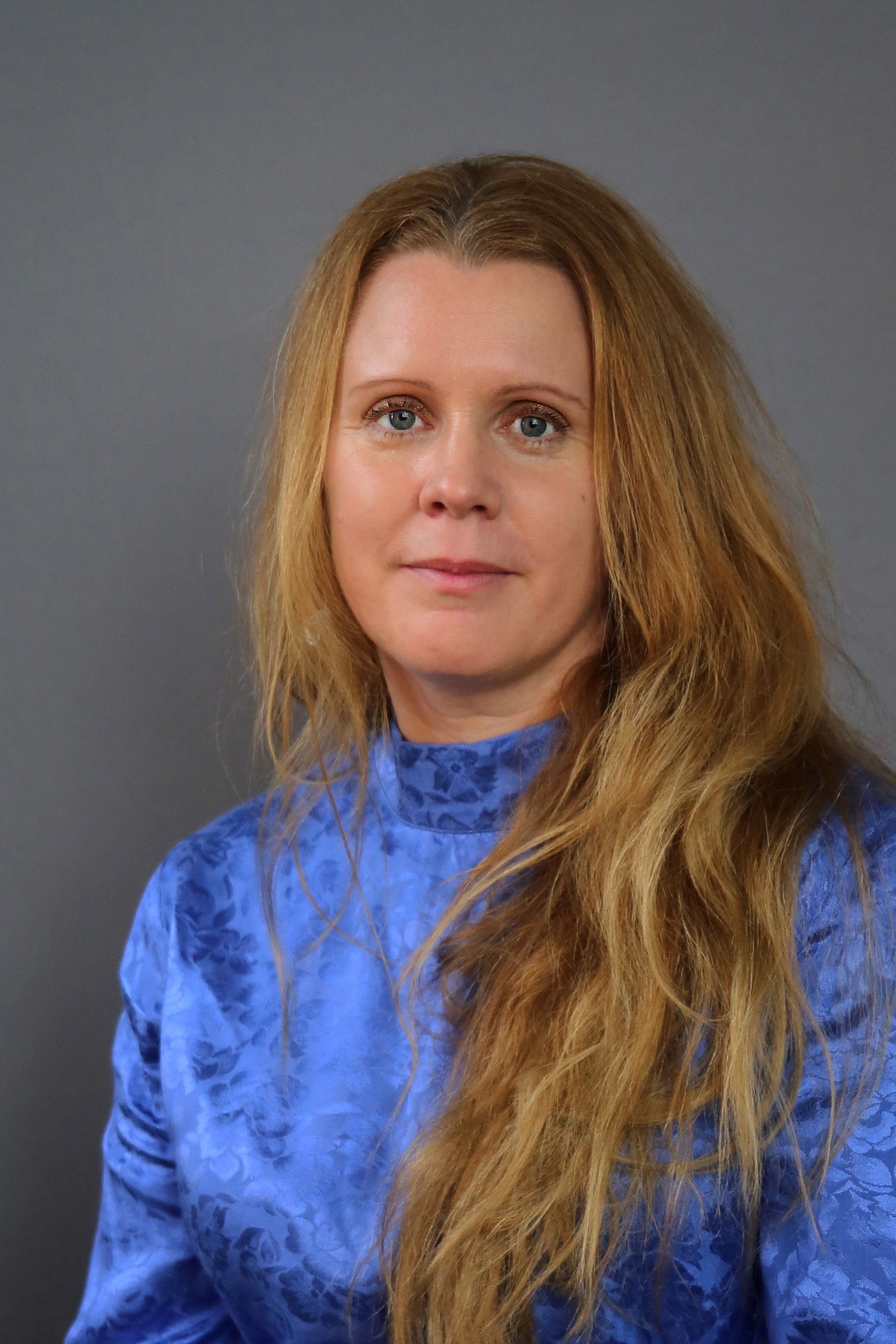 Kvinna med långt, blont hår och blåmönstrad topp sitter framför en grå bakgrund.