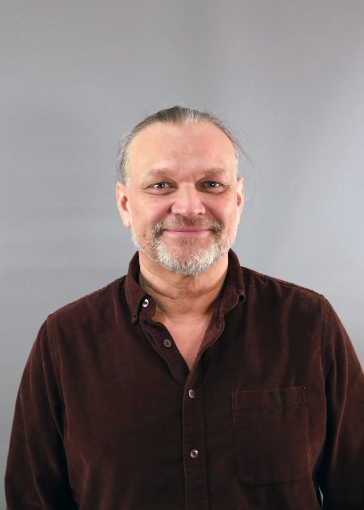 En medelålders man med skägg och brun skjorta ler mot en grå bakgrund.