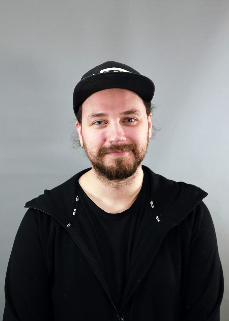 En man med skägg och keps ler lätt, iklädd svart huvtröja och t-shirt mot en grå bakgrund.