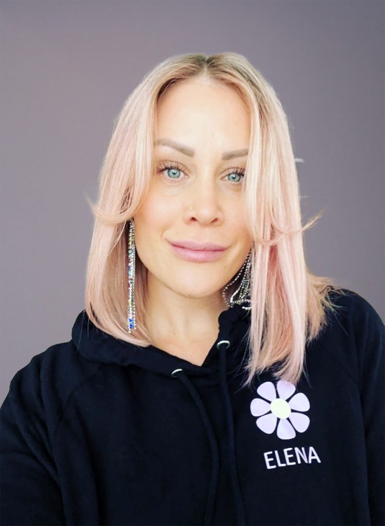 Kvinna med blont hår i svart huvtröja med texten "ELENA", ler mot kameran mot en enfärgad bakgrund.